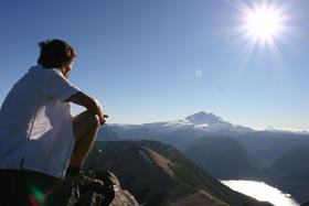Climber sitting on a summit looking across a lake and smaller mountains at a large snow covered peak in the distance under bright beaming sun in a gorgeous blue sky.  Breakthrough NPD can help you chart a course across the landscape of business to achieve the financial objectives symbolized the mountain and a happier future exemplified by the sun and sky in this beautiful place.